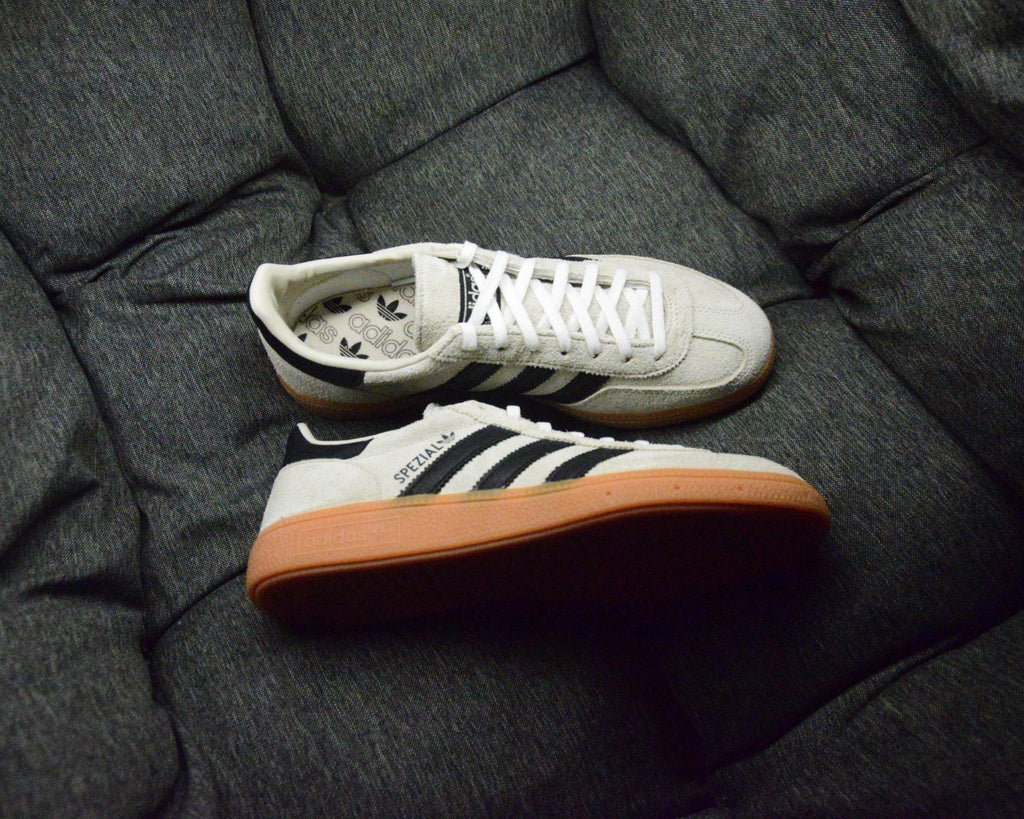 Adidas Handball Spezial Sneaker in Creme mit schwarzen Streifen und brauner Gummisohle auf dunklem Sofa